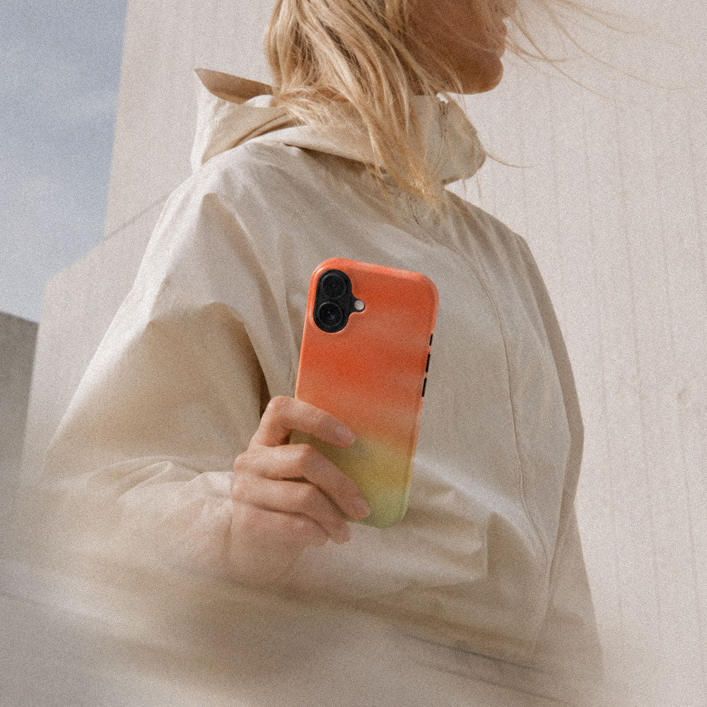 Kvinne i lys jakke som holder en iPhone med Koraldis mobildeksel i korall og gul gradient.