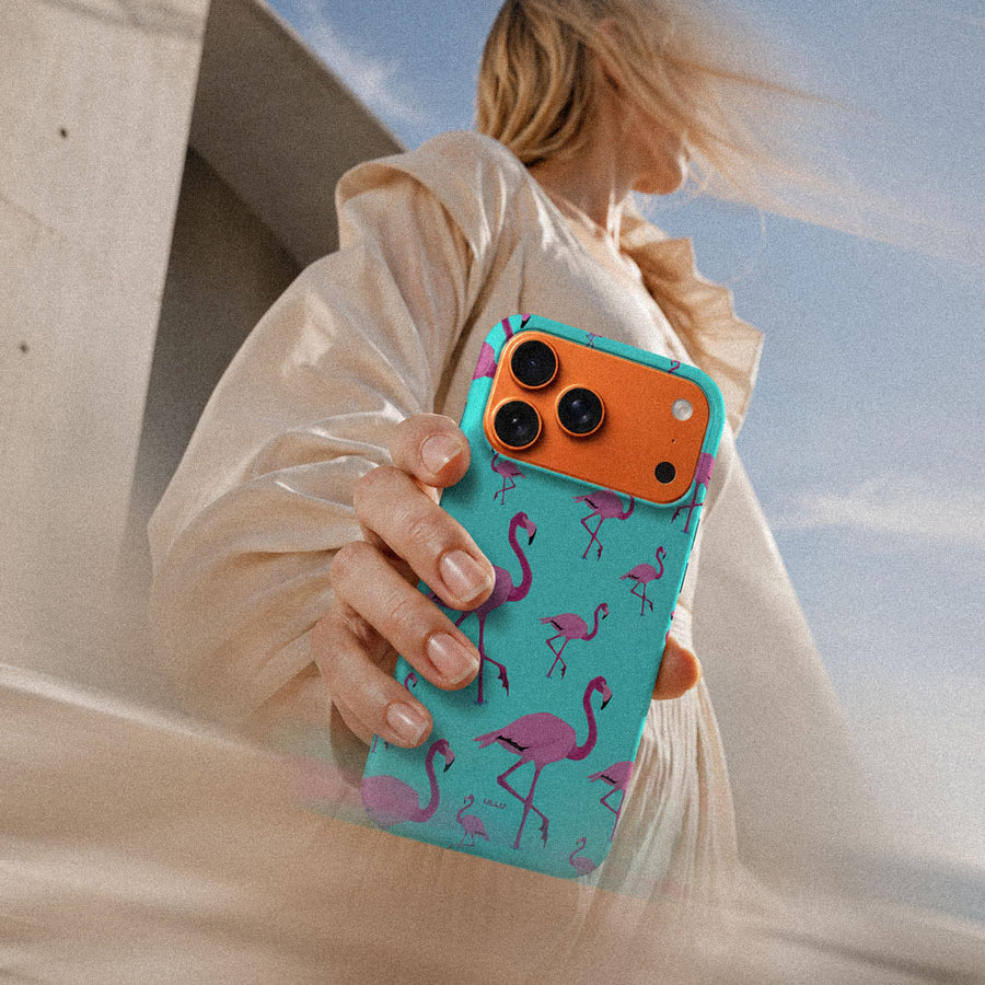 Kvinne i lys kjole som holder en iPhone 17 Pro Max med Flamingo mobildeksel og rosafargede flamingoer på turkis bakgrunn.
