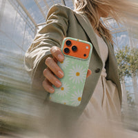Kvinne i grønn blazer som holder en iPhone 17 Pro Max med Kamille mobildeksel og hvite prestekrager på lys grønn bakgrunn.