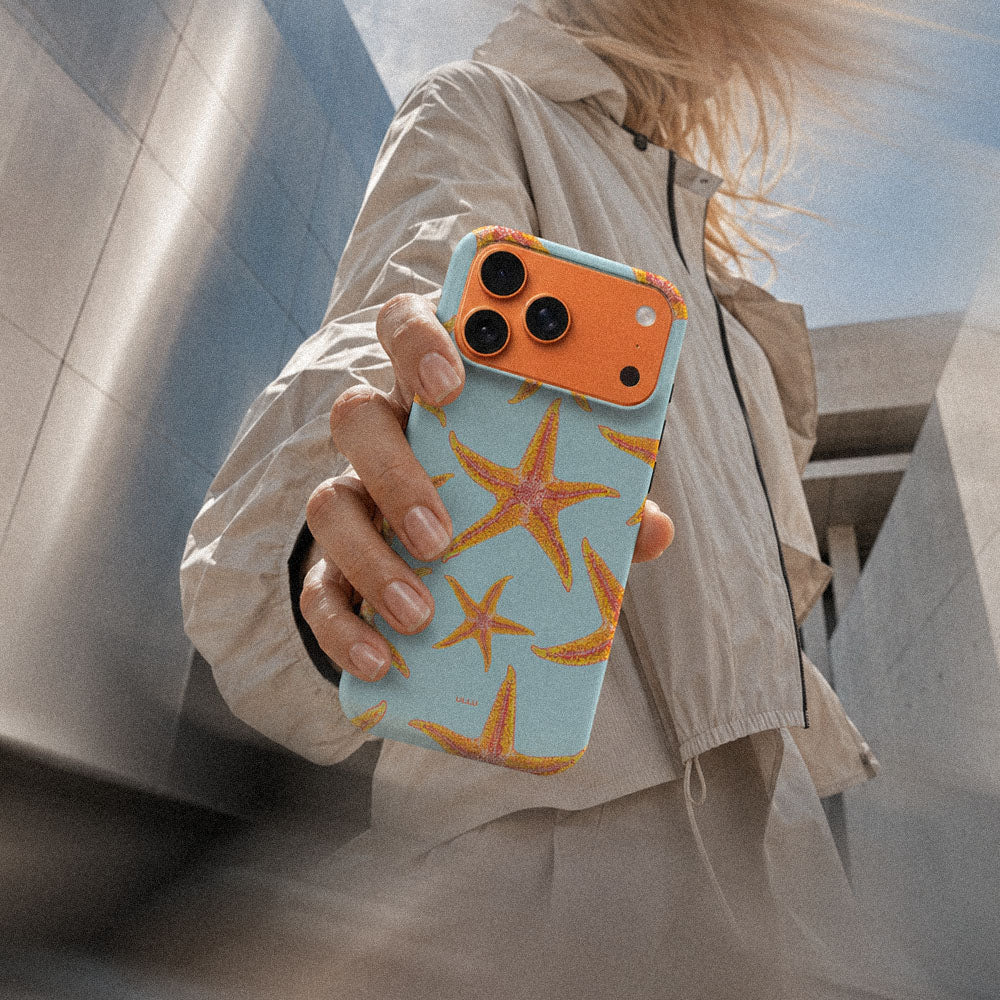 Kvinne i lys jakke som holder en iPhone 17 Pro Max med Sjøstjerner mobildeksel og oransje sjøstjerner på lys blå bakgrunn.