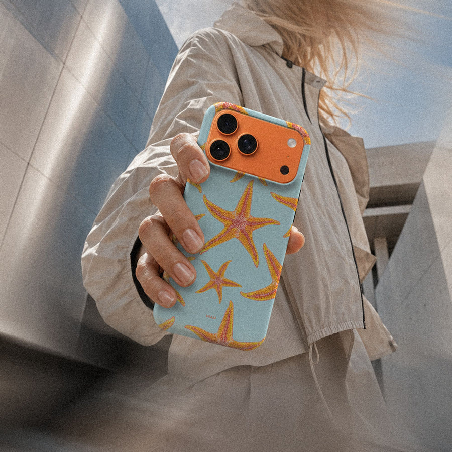 Kvinne i lys jakke som holder en iPhone 17 Pro Max med Sjøstjerner mobildeksel og oransje sjøstjerner på lys blå bakgrunn.